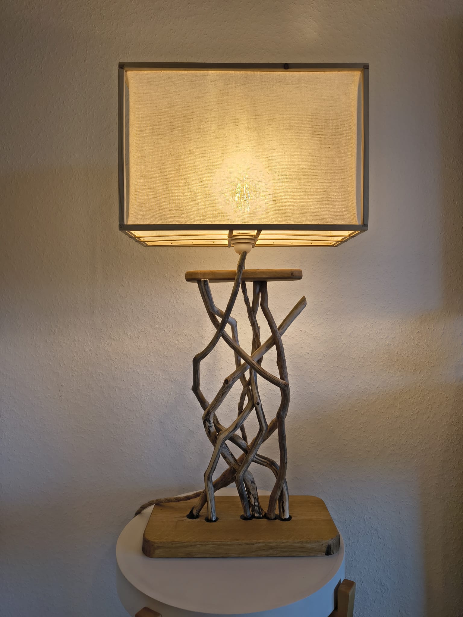 Lampe Racines allumée dans la pénombre, pied en bois flotté noué et abat-jour en lin